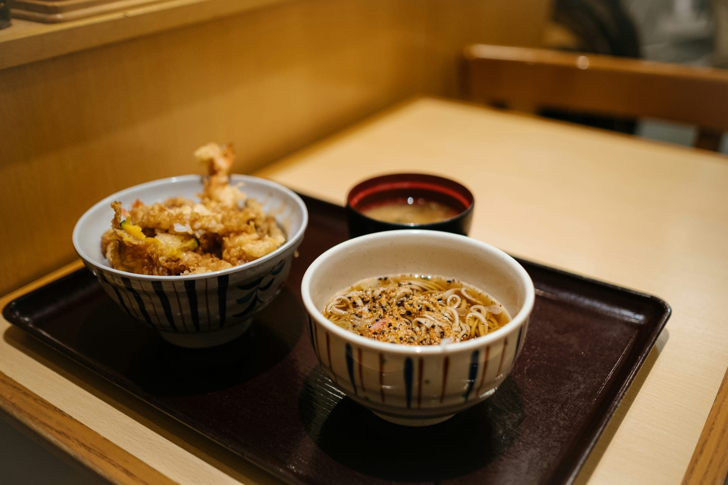 木のカウンターに置かれた漆の盆。天丼と、温かいつゆに入った小丼のそばと、赤い椀の味噌汁が載っている。よその店の卓で、うちの器でも品書きでもない。